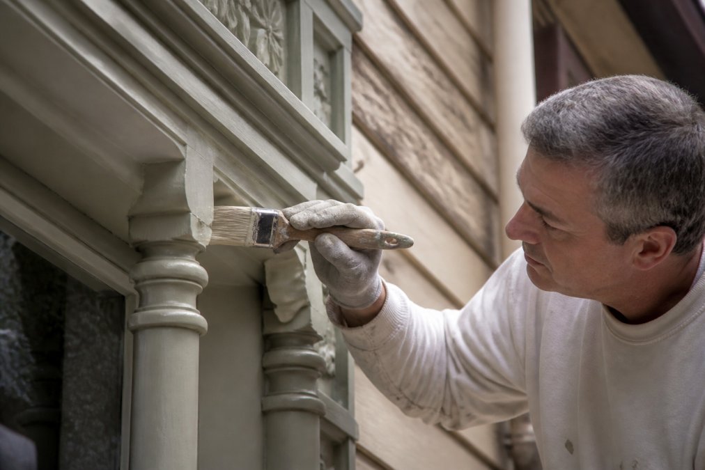 Professional painter carefully applying period-accurate paint to ornate heritage timber trim, showing meticulous surface preparation and brushwork on decorative detailing with natural workshop lighting