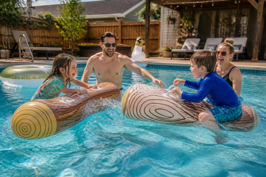 Family laughing and splashing while playing with inflatable pool toys on battle log rafts in a sunlit backyard pool.