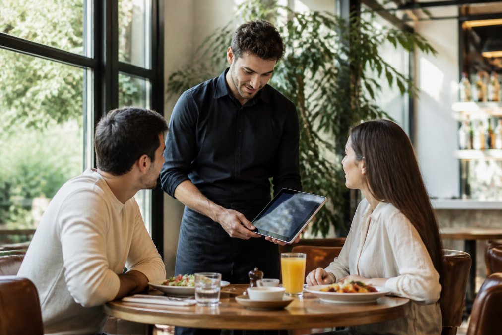 Camarero presentando menú digital en tablet a comensales sentados en mesa de restaurante moderno, con platos y bebidas sobre la mesa, iluminación natural cálida