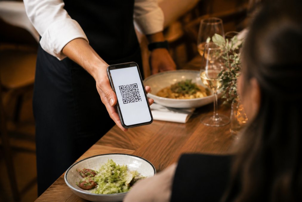 Un camarero sostiene un smartphone mostrando un menú digital con código QR sobre una mesa de madera, con platos de comida y copas desenfocadas al fondo, luz cálida de restaurante
