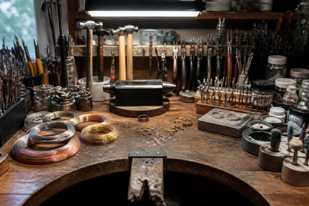 Professional jeweler's workbench with organized metal smithing tools, wire coils, finished metal components, and polishing supplies arranged under bright overhead lighting
