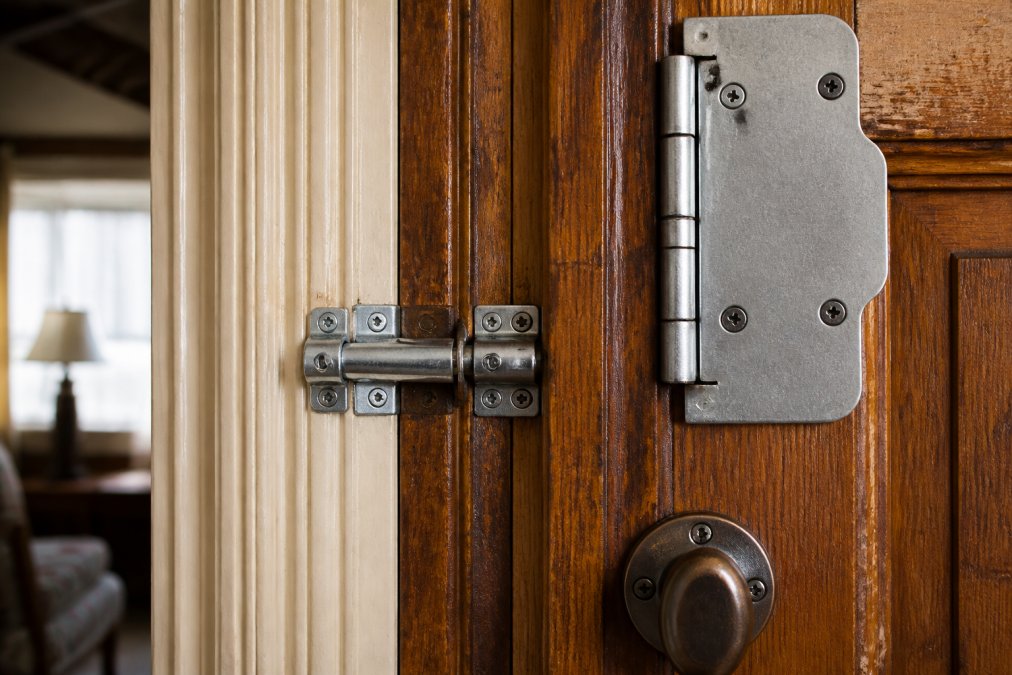 Close-up of galvanized steel security bolt and hinge shield installed on vintage wooden door frame, showing how metal reinforcement integrates with original wood trim and period hardware without visible exterior changes