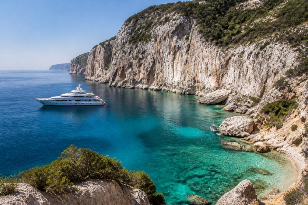 White superyacht anchored in a turquoise Mediterranean yacht cruising cove beneath towering limestone cliffs.