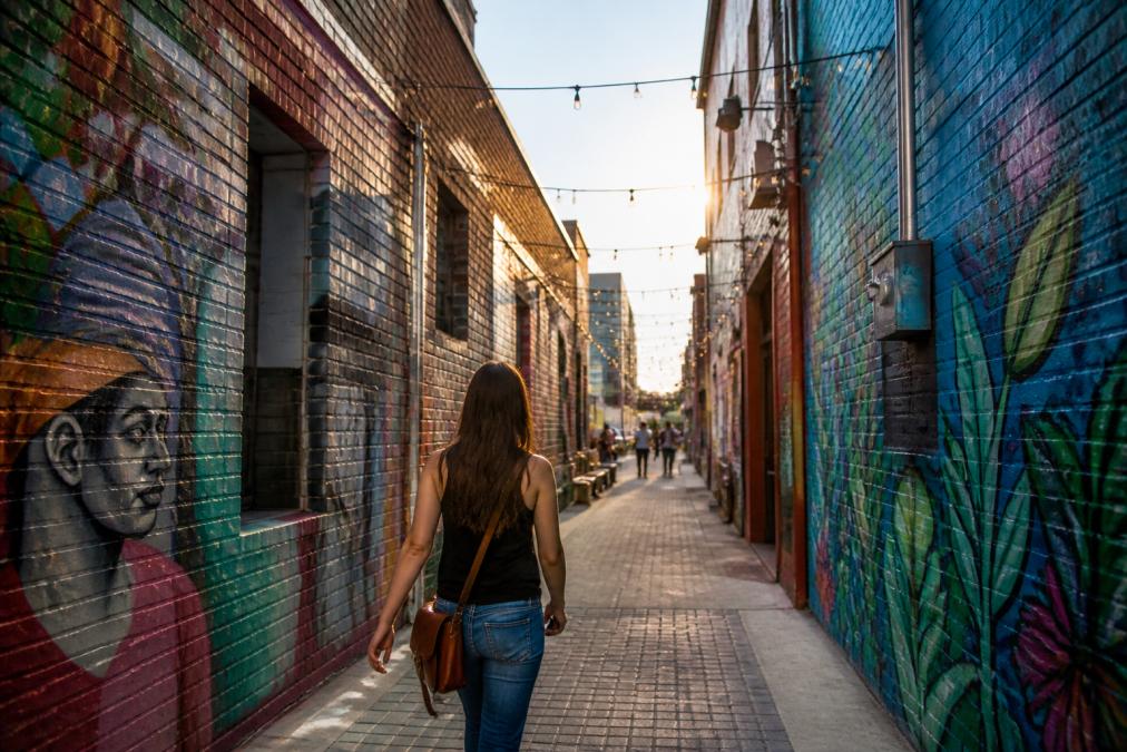 A person walking through a narrow, art-filled alleyway in Omaha with colorful murals and string lights overhead, late afternoon light
