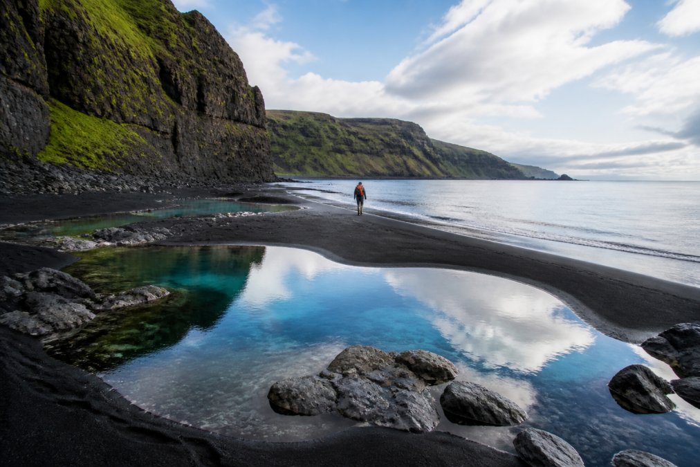 A lone hiker standing on a dramatic black sand beach with crystal-clear turquoise pools reflecting the sky, surrounded by towering rugged cliffs and untouched wilderness
