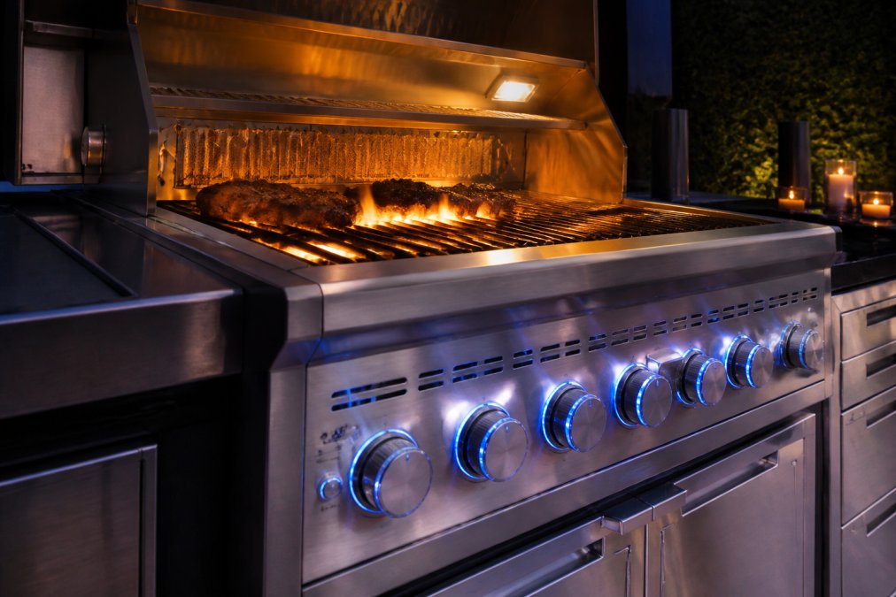 Close-up of a premium stainless steel grill with illuminated LED control knobs and dual infrared burners glowing during evening outdoor cooking, with flames visible through the grate