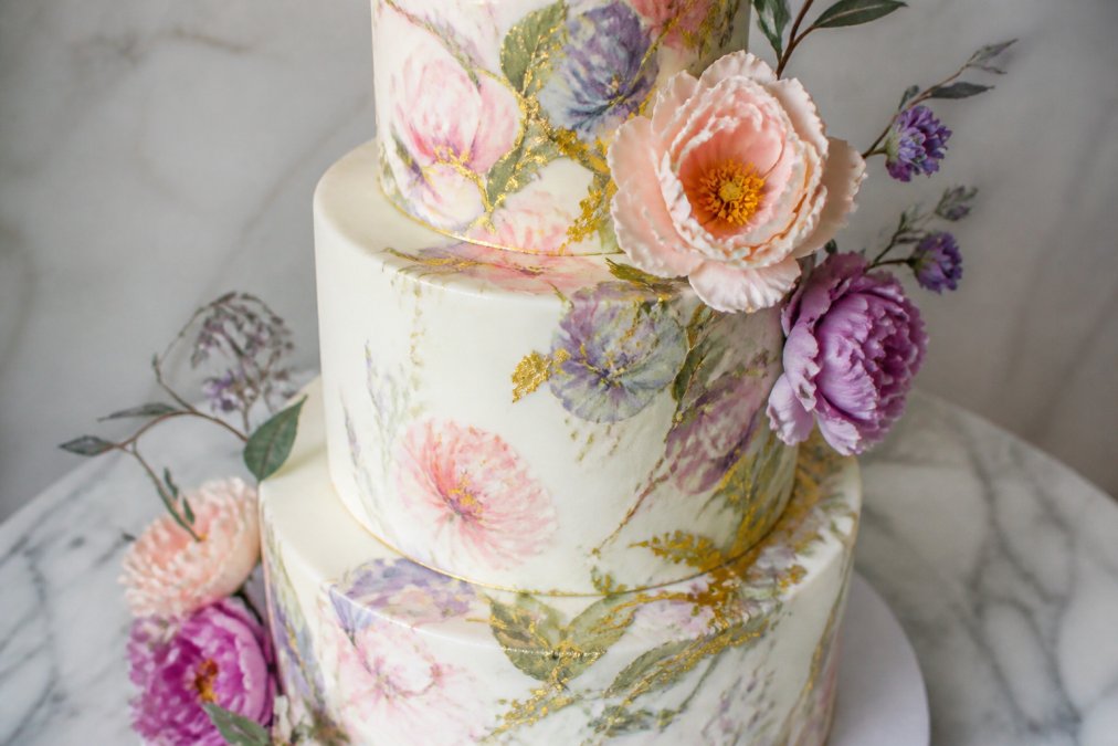 Close-up of an elegantly decorated multi-tiered birthday cake with hand-painted watercolor florals, edible gold leaf accents, and delicate sugar flowers displayed on white marble with soft natural lighting