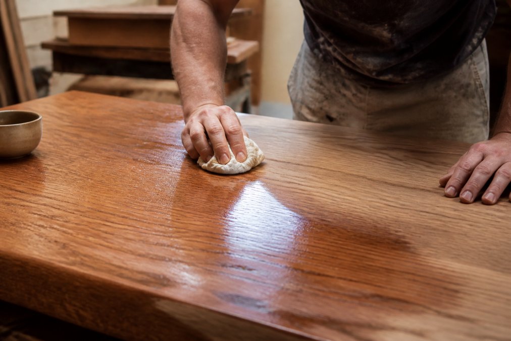 Artisan's hands carefully applying hand-rubbed lacquer finish to solid oak timber surface in natural workshop lighting, showing wood grain detail and finishing technique