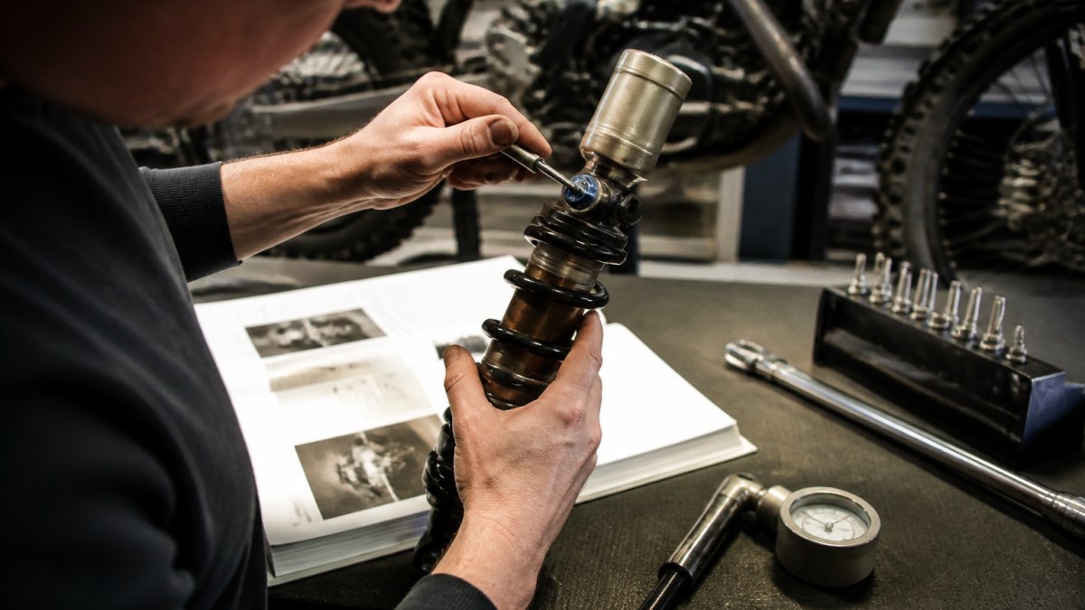 Close-up van motorrijder die aan compressie- en rebound-instellingen van een offroad-veringsysteem werkt met fijne instellingstools, terwijl een handleiding en meetinstrumenten op de werkbank liggen