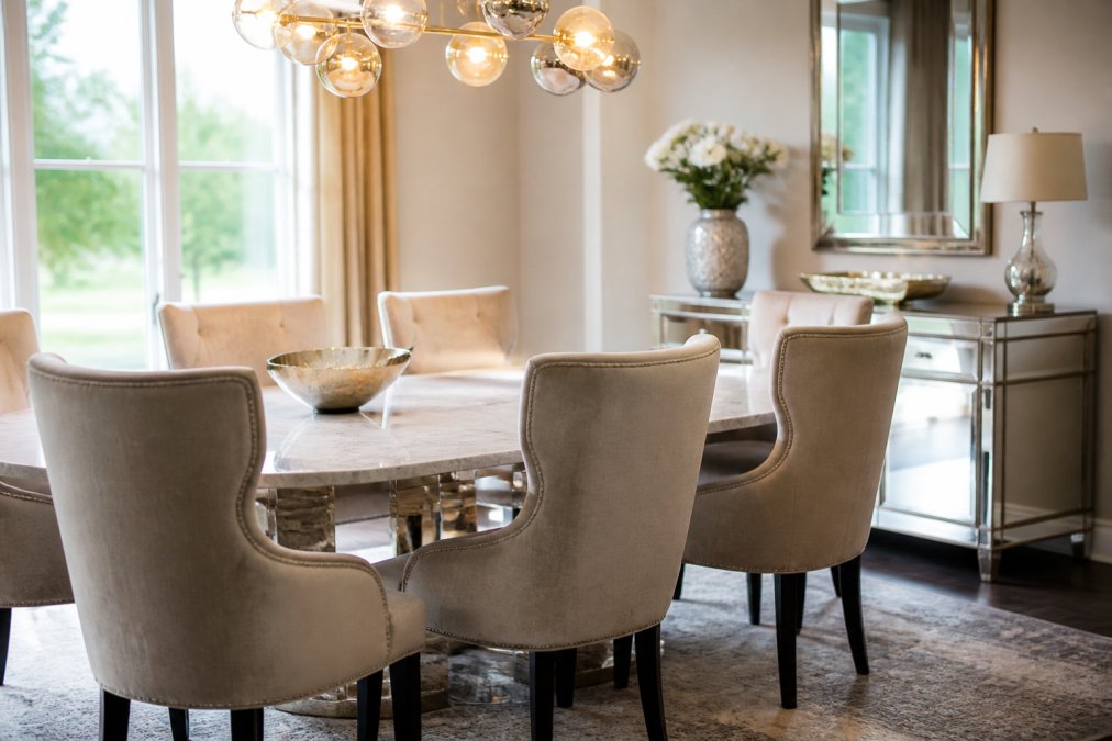 Modern living room with beige velvet wingback dining chairs positioned around a faux marble table, mirrored console table reflecting soft natural light from large windows, contemporary pendant lighting overhead creating warm ambiance