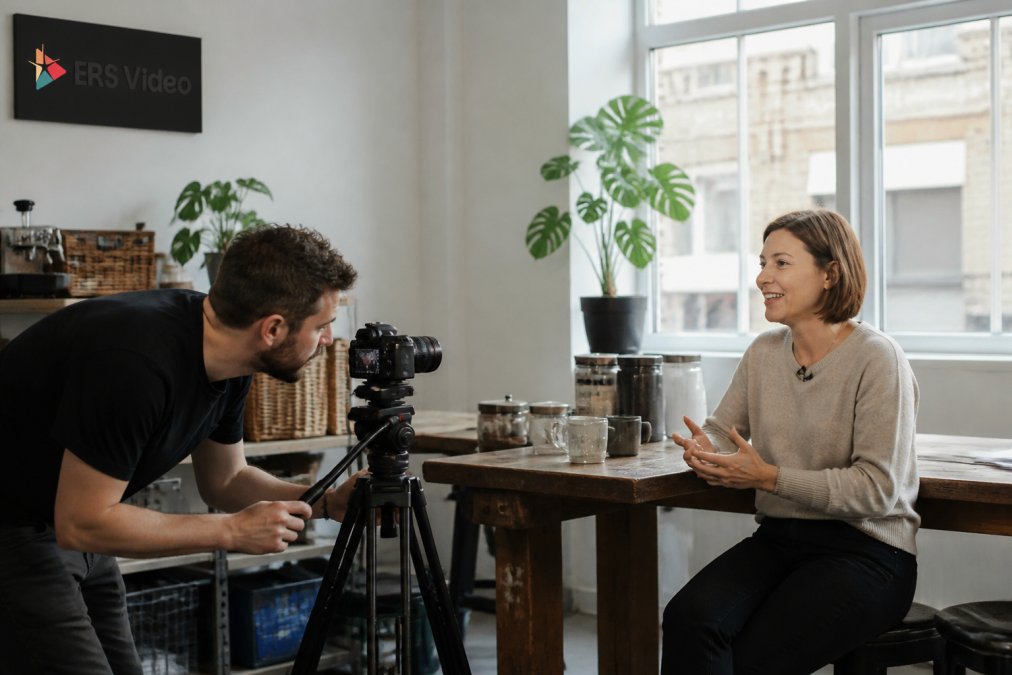 A small business owner being interviewed on camera in their workplace, with a videographer adjusting a tripod in the foreground and natural light from a window