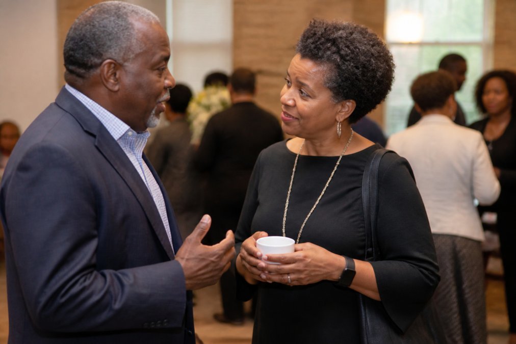 Two professionals in conversation at a community event, one holding a coffee cup, engaged in meaningful dialogue with warm lighting and community members visible in the background