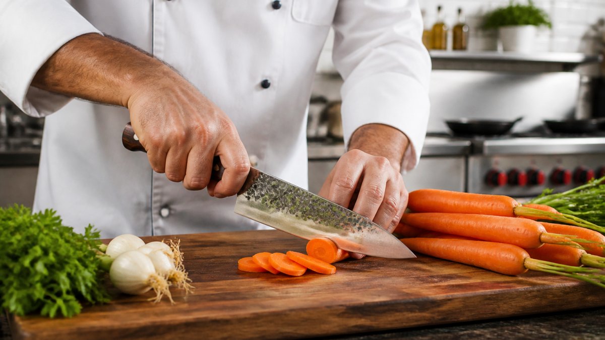 Professional chef demonstrating proper grip on a handcrafted custom chef knife while preparing fresh vegetables on a wooden cutting board in bright kitchen lighting