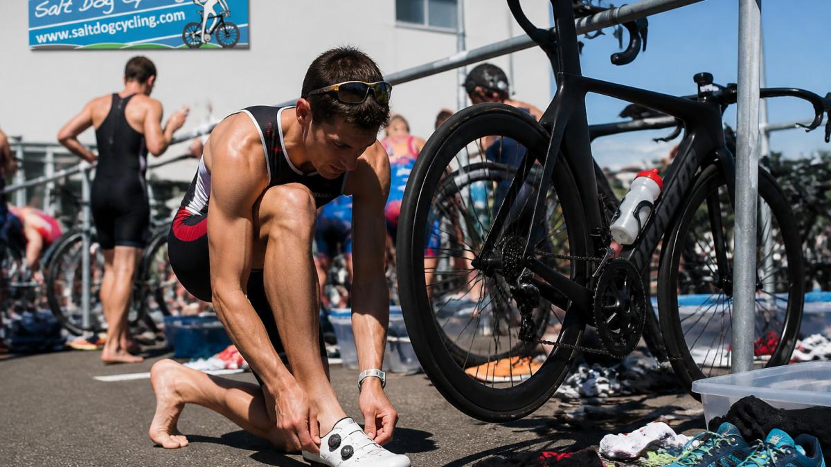 Triathlete putting on cycling shoes in a race transition area with bikes racked nearby