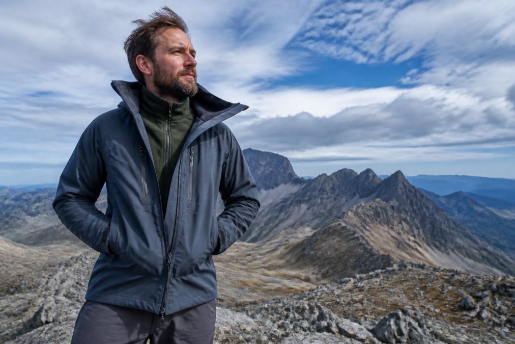 A hiker standing at a mountain summit wearing a shell jacket over a visible mid-layer fleece, wind pressing against the fabric, dramatic rocky ridgeline and cloud-streaked sky in the background