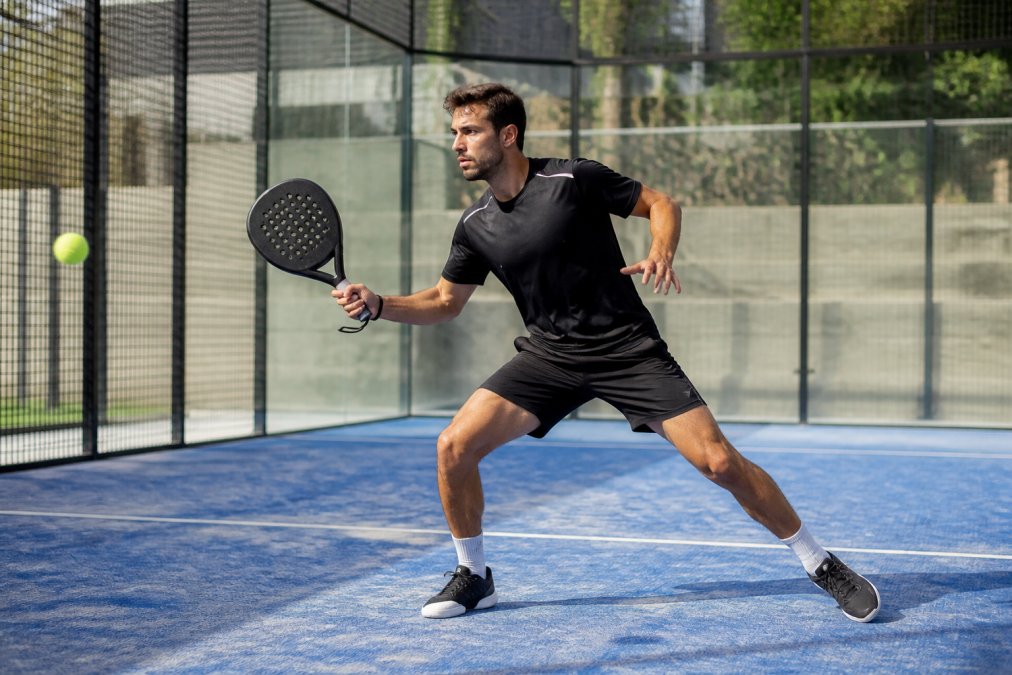 Male padel player mid-rally on outdoor court wearing technical black shorts and moisture-wicking shirt, demonstrating explosive lateral movement and agility