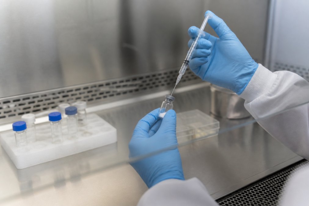 Hands of a researcher in sterile gloves carefully drawing sterile reconstitution solution with a syringe, about to inject it into a vial of lyophilized peptide powder in a clean laboratory hood with proper lighting