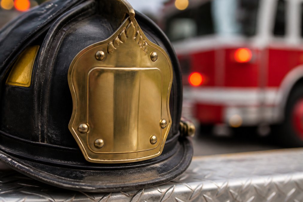 Close-up of a firefighter's leather helmet with a polished brass front holder mounted on the front, showing the rivets and engraved detail, with a fire truck blurred in the background