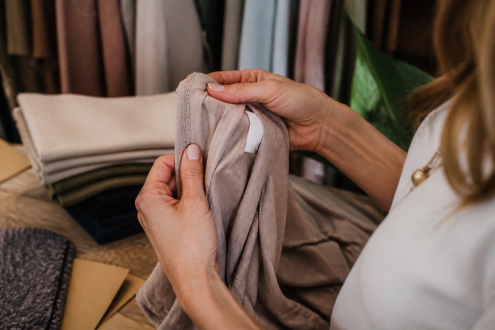 Close-up of a woman's hands examining the interior seam and fabric label of a garment under warm boutique lighting, fingers tracing the stitching detail along the seam allowance