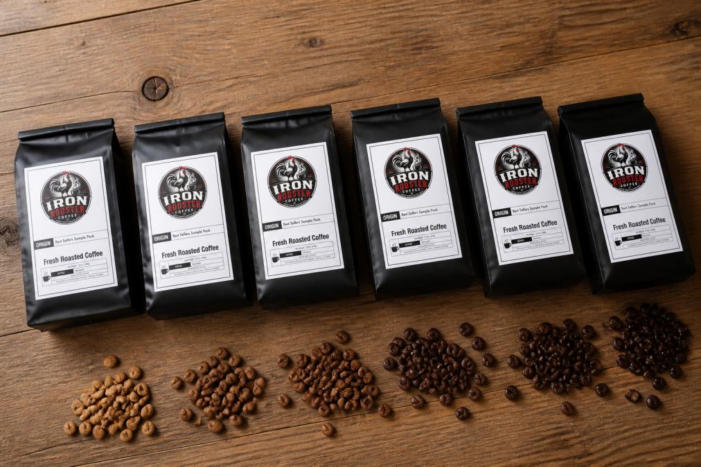 Flat lay of six small coffee bags with different roast levels arranged on a wooden surface, showing color progression from light to dark roasts with fresh beans scattered nearby