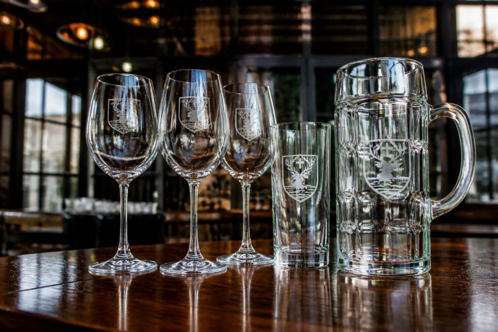 Close-up of laser-engraved wine glasses and beer steins with corporate logos displayed on a polished wooden table, showing precision etching detail and reflective quality of premium glass