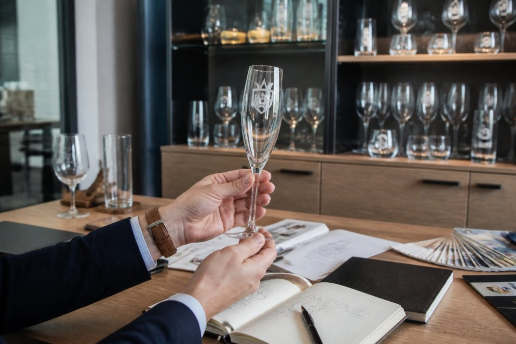Professional hands holding a champagne flute with custom branding, positioned against a corporate office background with branded glassware collection visible on shelves
