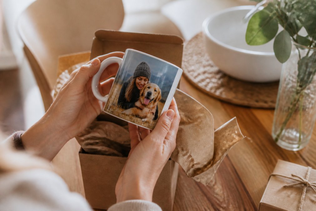 A person's hands holding a freshly unwrapped ceramic mug with a personalized photo print, set on a kitchen table with soft natural light and a gift box nearby