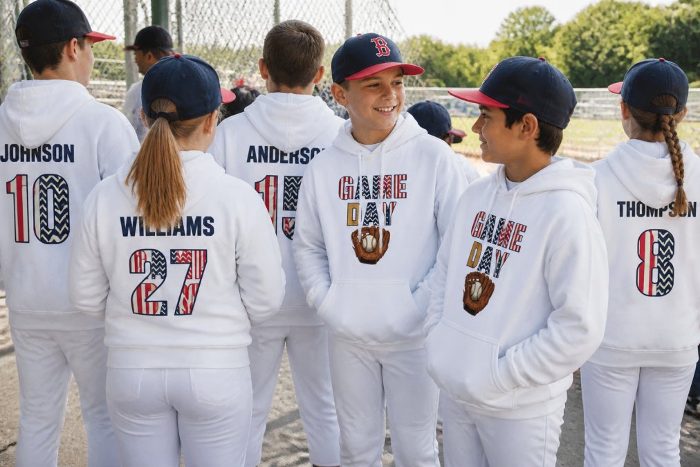Youth baseball and softball team wearing matching custom hoodies and personalized jerseys with names and numbers visible, standing together on sidelines in natural daylight showing team unity and spirit