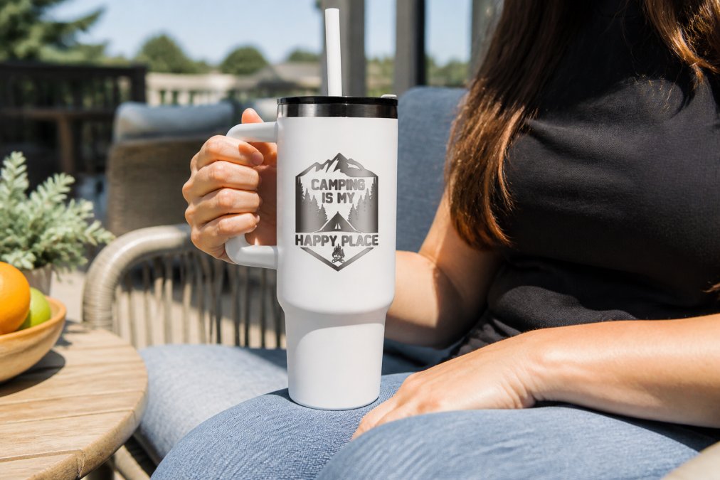 Person holding a custom laser-engraved stainless steel tumbler with personalized design while enjoying a cold beverage on a bright outdoor patio, morning sunlight illuminating the matte finish and crisp engraving detail