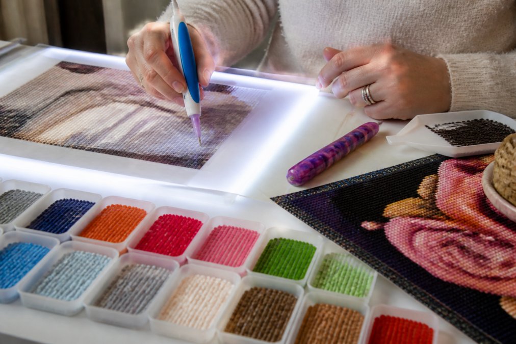Crafter's hands holding an electric diamond painting pen positioned over a canvas placed on an illuminated light pad, with organized colorful drill trays arranged nearby and a partially completed sparkled diamond painting visible in the corner