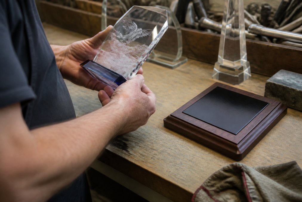 Close-up of a craftsman's hands inspecting a laser-engraved crystal award and a walnut plaque on a workbench, fine detail visible in the etching under bright workshop lighting