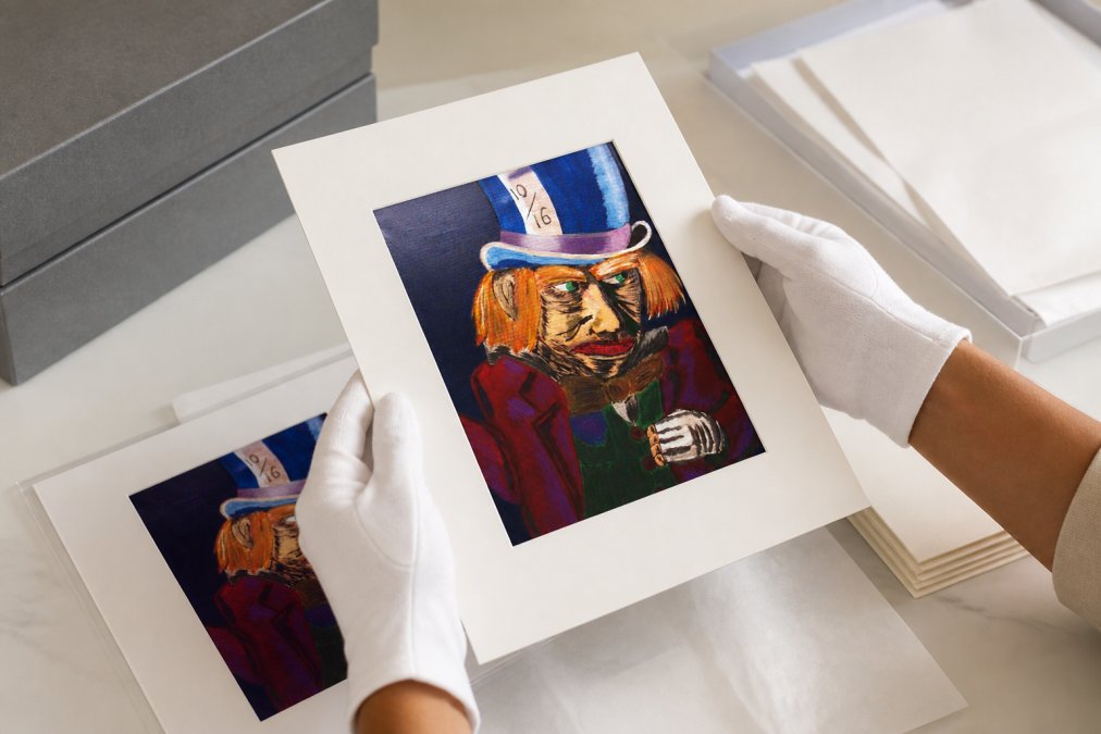 Hands carefully handling a fine art print with archival matte finish, showing proper grip technique while wearing cotton gloves, with acid-free materials and storage boxes visible on a clean workspace