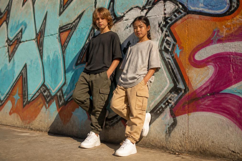 Two kids wearing oversized graphic tees and cargo joggers in an urban outdoor setting, styled casually with white sneakers, leaning against a graffiti-covered wall in warm afternoon light, looking relaxed and confident
