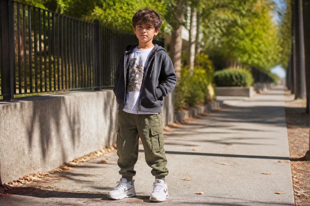A child wearing a layered streetwear outfit with a zip-up hoodie over a graphic tee and cuffed cargo joggers, standing on a sunlit footpath with a relaxed, confident pose, sneakers visible, natural urban background