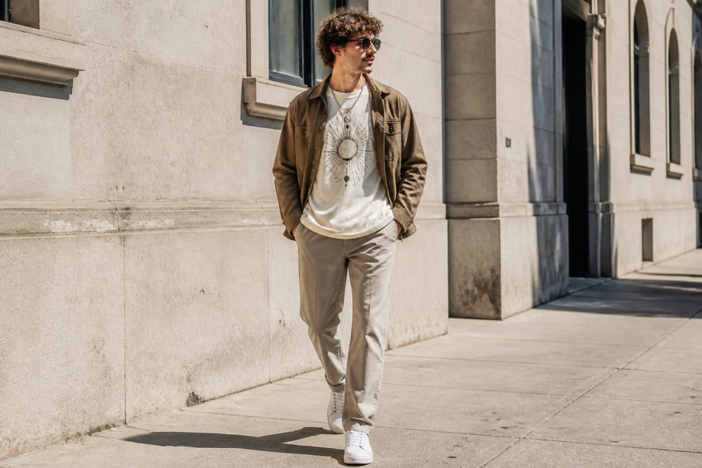 A person layering an oversized minimalist graphic tee under an open earth-toned jacket, paired with straight-leg pants and clean white sneakers, photographed outdoors on a sunny urban sidewalk with natural daylight