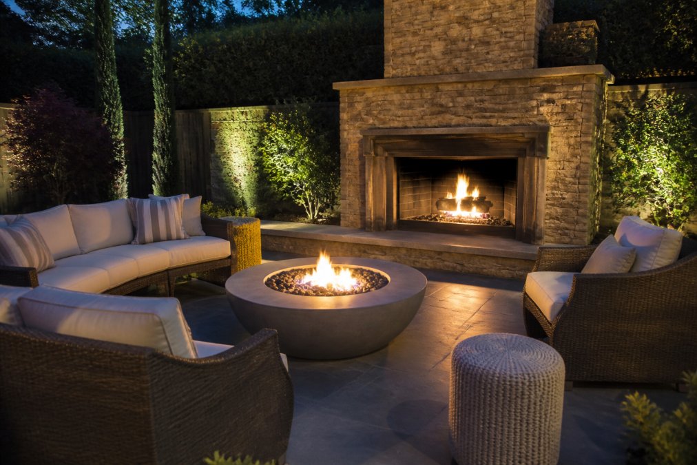 Modern backyard patio at dusk with a glowing gas fireplace, comfortable seating arranged around the fire bowl, warm ambient lighting creating an inviting outdoor living space with landscaping