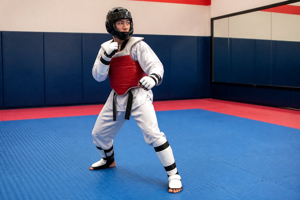 A martial artist in a crisp white dobok wearing full taekwondo sparring gear, including a red chest protector, black headgear, gloves, and shin guards, standing in a fighting stance on a bright dojo floor