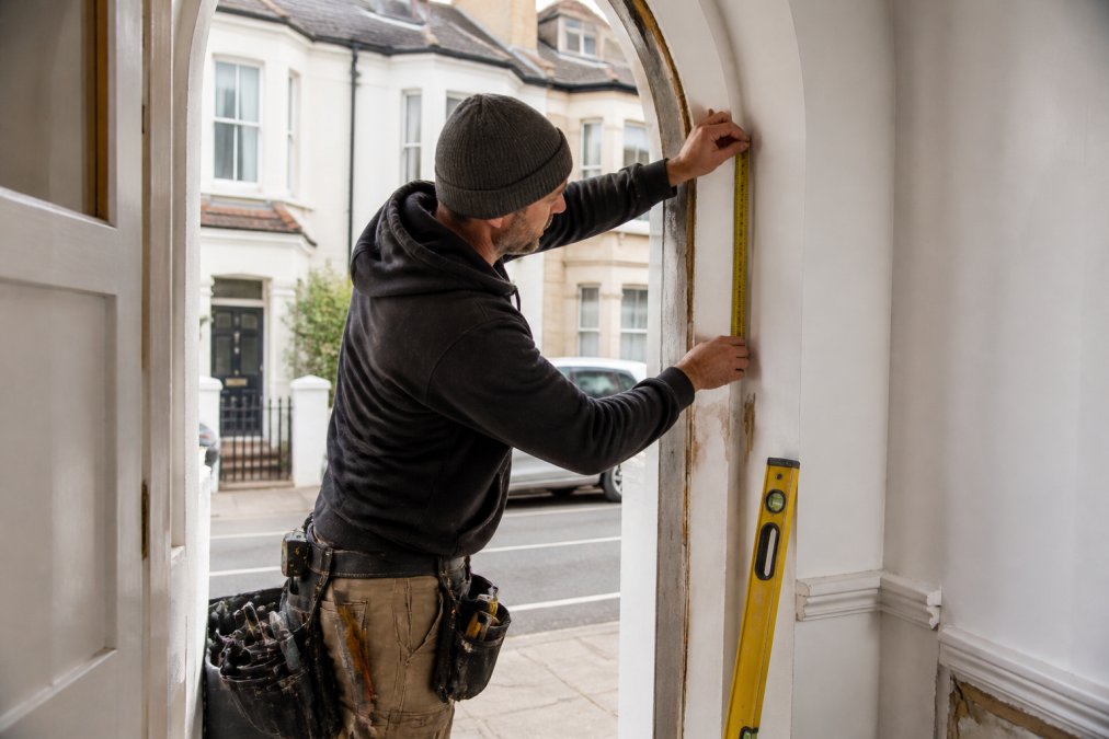 Skilled joiner or craftsperson in workshop attire measuring a non-standard Victorian terrace opening with precision tape measure and spirit level, taking detailed measurements of an arched or irregular doorway