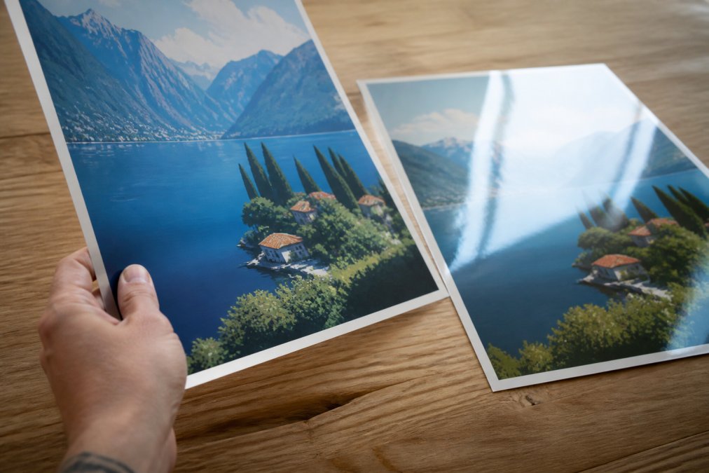 Close-up of a hand holding a premium matte travel poster at an angle, showing the non-reflective surface and rich colour depth, with a thin glossy print lying flat on a wooden table beside it showing visible light reflection