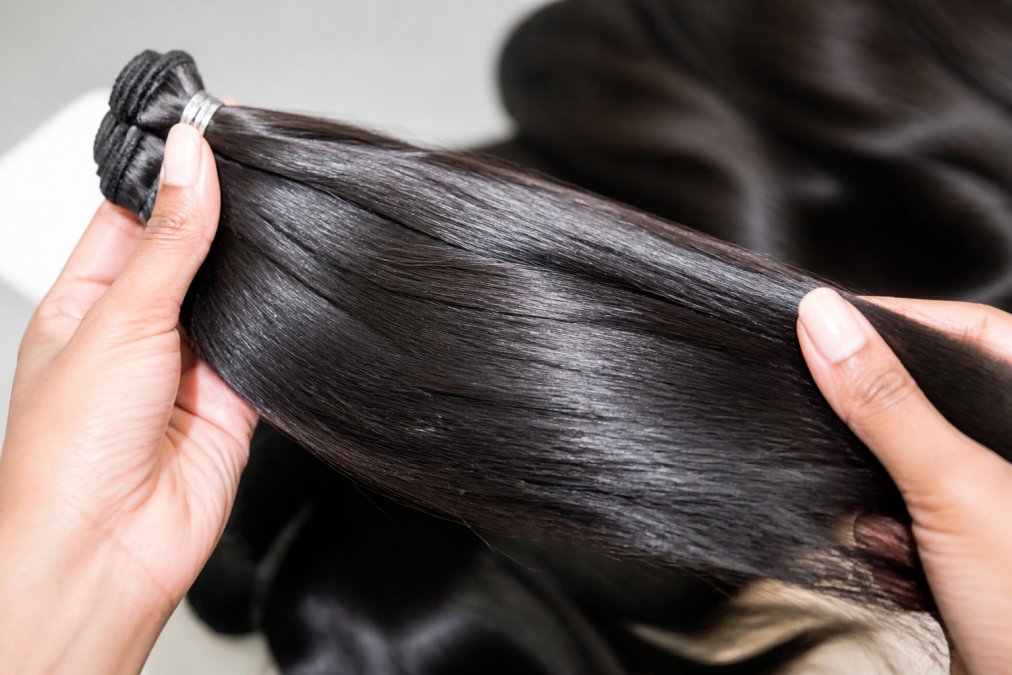 Close-up of a person's hands holding a section of natural dark virgin hair weft against a soft neutral background, showing the cuticle-aligned texture and natural sheen