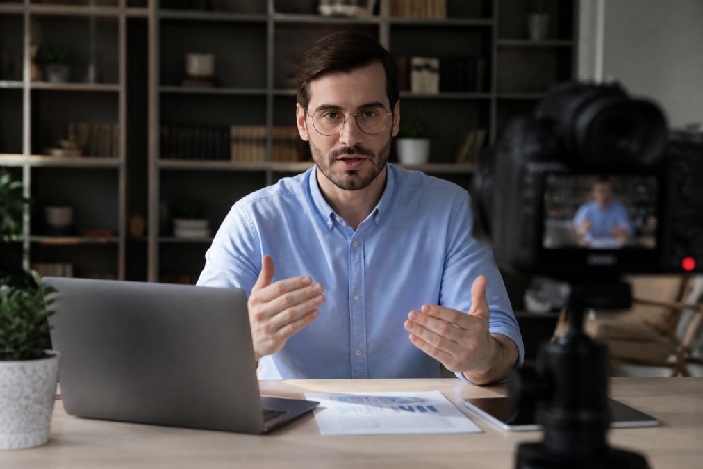 Business professional presenting to camera during live webcast, with engagement metrics and real-time audience interaction visible on screens showing live polls, chat messages, and participant counts