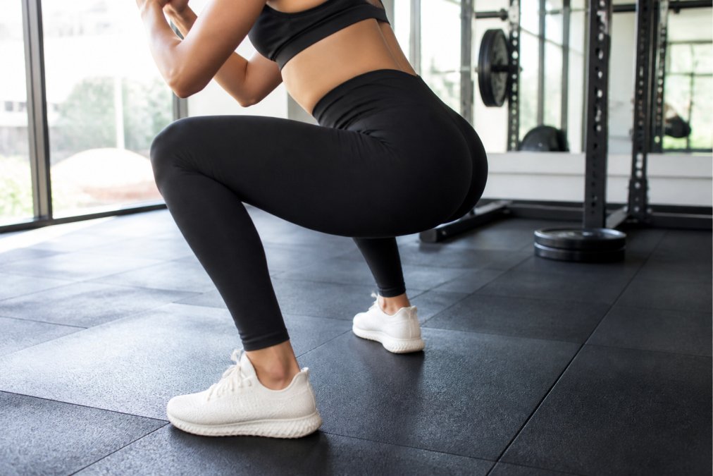 Close-up of a woman in high-waisted black workout leggings performing a deep squat in a bright, modern gym with large windows, fabric visibly stretching without sheering