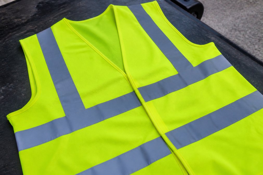 High Visibility Roadside Safety Equipment Guide Close-up of a high-visibility vest with bright fluorescent yellow-green material and silver retro-reflective strips across the shoulders and torso, laid flat on a dark surface in natural daylight