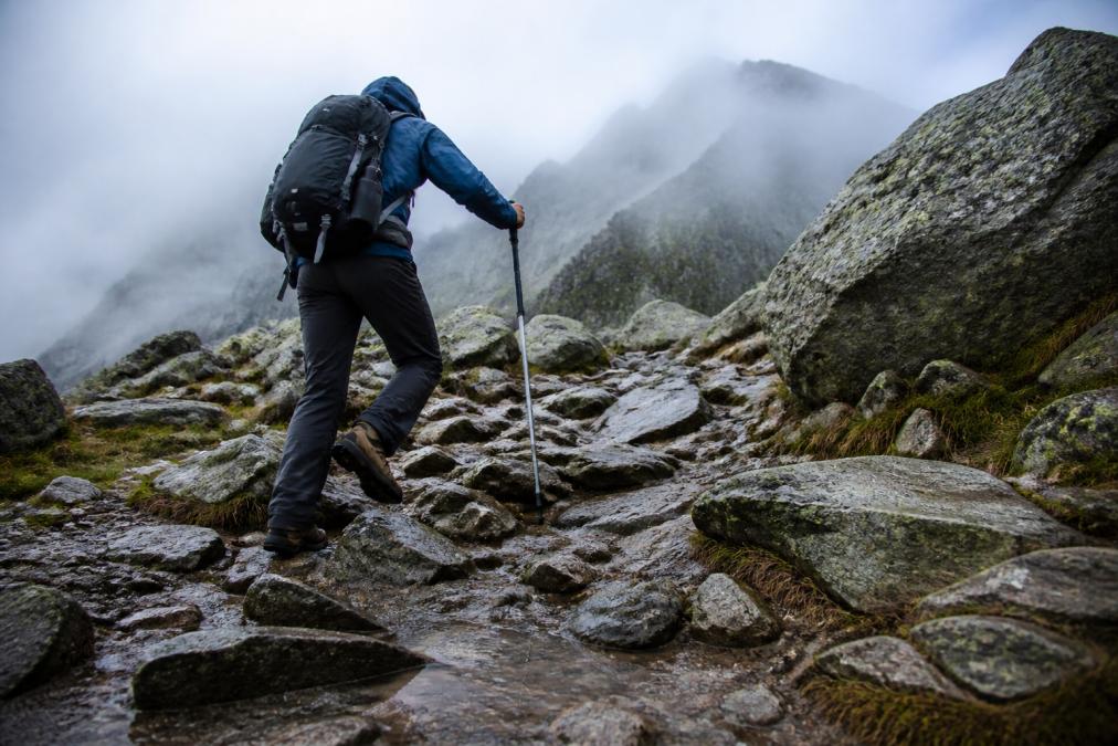 A hiker wearing sturdy boots navigating a rocky, wet mountain trail with a loaded backpack, stepping carefully over uneven stones