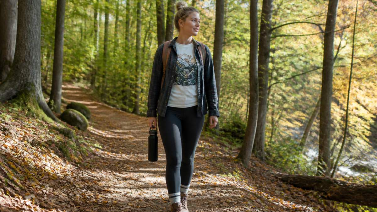 A person in a soft cotton graphic tee and light layers walking a flat woodland trail on a mild fall morning, with a small daypack and a water bottle, natural light through the trees