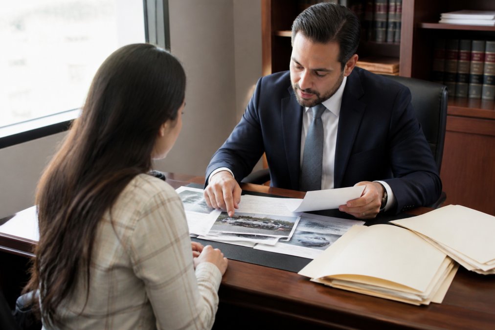 Personal injury lawyer reviewing accident scene photographs and medical records with client seated across desk in professional office with natural window lighting