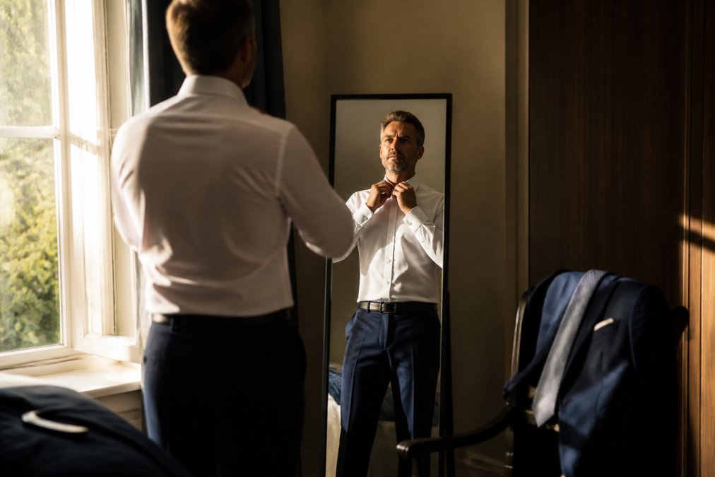Professional in smart business attire adjusting collar and checking appearance in mirror, natural morning light streaming through window, wardrobe pieces visible on chair nearby