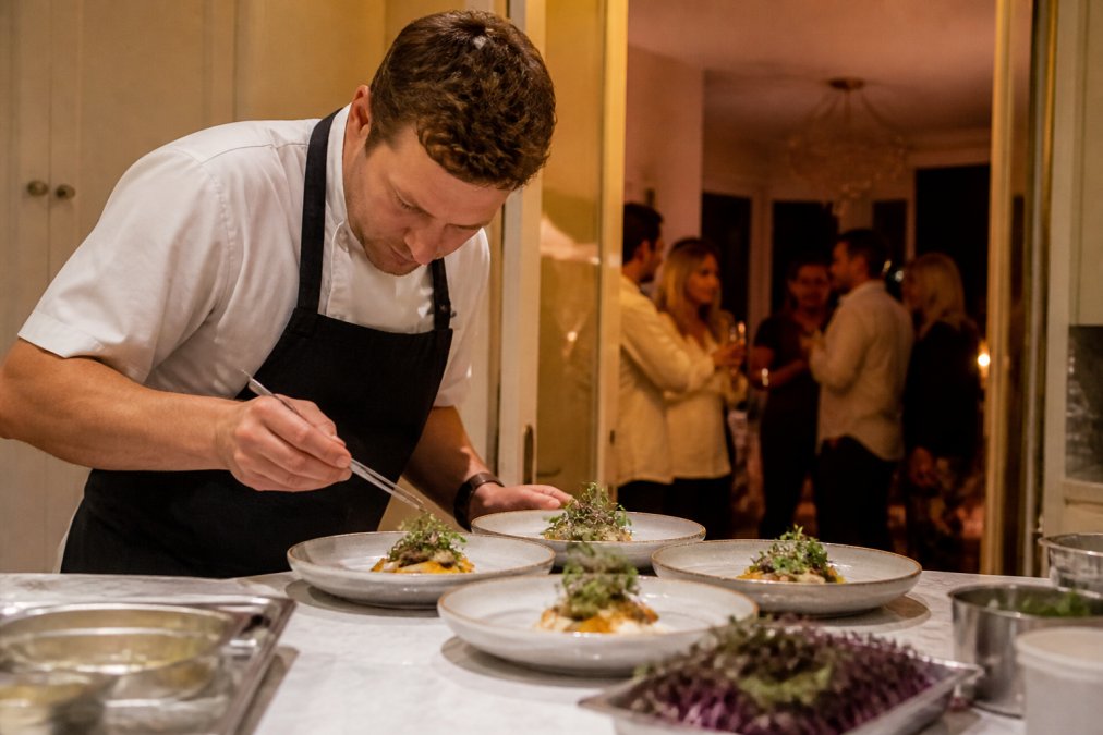 A private chef plating a fine dining dish on a kitchen island in a home, guests visible in soft focus through a doorway in warm evening light