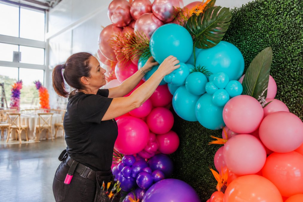 Professional balloon artist in colorful event space carefully arranging a custom balloon arch with organic backdrop, hands positioning latex balloons and decorative elements, natural venue lighting highlighting the vibrant colors