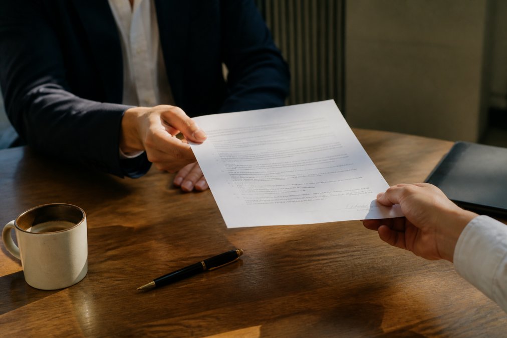 Close-up of two hands exchanging a signed contract document on a wooden desk, with a pen and a coffee cup visible in the background